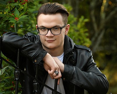 senior picture of louisville kentucky graduate. boy sitting on chair in black leather jacket and black rimmed glasses with black hair.
