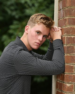 senior picture of louisville kentucky graduate. boy with blond hair and grey collared shirt leaning on brick wall