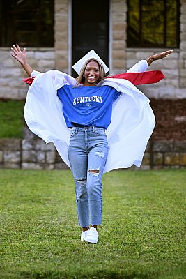 senior photography of louisville kentucky graduate. teenage girl in white graduation robe and blue "kentucky" t-shirt jumping in the air