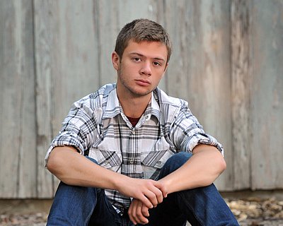 senior photography of louisville kentucky graduate. teenage boy in white and black plaid shirt, sitting, posing in front of wood background