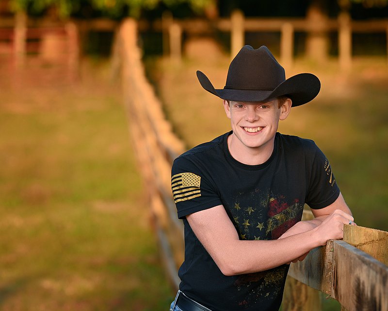 senior picture of louisville kentucky graduate. boy in black shirt and black cowboy hat leaning on fence outside